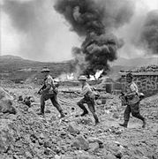 Men of 1st Battalion, The Duke of Wellington's Regiment, advance past a burning fuel store on Pantelleria during Operation Corkscrew