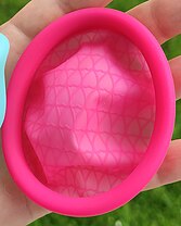 A menstrual disc/ring, made of silicone, bright pink. The rim has a round cross-section and is about 1cm thick; it forms an oval. The bowl is a crumpled, slightly-translucent silicone membrane, with a latticework of slightly-raised ridges in the shape of a tesselation of hearts.