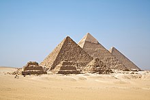 Frontale Farbfotografie in der Obersicht von drei hintereinander stehenden Pyramiden in der Wüste. Vor der ersten Pyramide befinden sich drei kleine Pyramidenruinen. Links laufen Menschen mit Pferden.