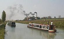 A straight section of canal going into the distance, with two passing narrow boats in the foreground, with a large brick building and tall chimney in the middle-distance on the right bank of the canal. Smoke is billowing from the chimney, blowing across the canal to the left.