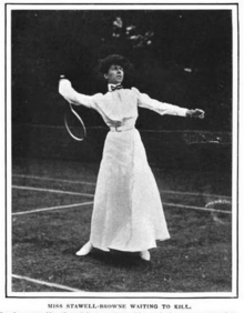 A white woman standing on a tennis court in tennis whites, including an ankle-length skirt, holding a tennis racquet behind her head in a position of readiness
