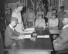 Men in uniform sit at a large wooden table. One is signing a document.