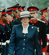 Thatcher in a blue suit and hat, walking in front of troops