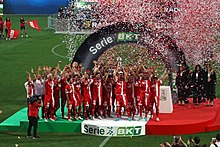 Football players celebrating. The team's captain lifts a cup. Red and white confetti is shot in the background.