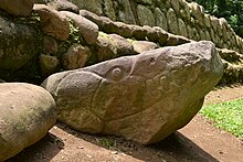 A roughly triangular boulder with the point inclined to the right. It has been carved in low relief to outline the jaws, teeth and eyes of a crocodile, with the point of the boulder representing the snout. A stonework structure rises behind the sculpture, which faces towards a grassy area to the right