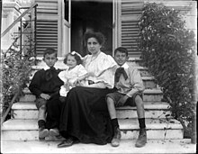 A woman and three children sitting on steps outside a door surrounded by bushes
