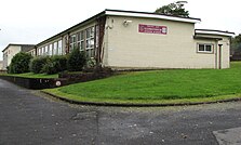 A picture depicting a side view of Llangennech Junior School. There is a red sign with white text containing "Ysgol Iau Llangennech Junior School" on a red background. There is also a grass verge.