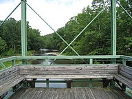 A detail of the wooden seat and observation platform at the center of the green Whipple truss bridge facing out over a river