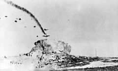 A blurry black and white photograph of two aircraft with numerous parachutes descending from them. One aircraft is trailing smoke and losing height