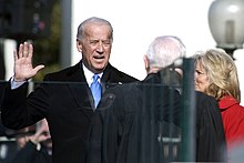 Joe Biden raising his right hand in front of a man in a robe, standing beside his wife