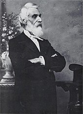 a man with a white beard and hair faces right, with his arms folded. He is dressed in a suit of a sort worn in the late 19th century.