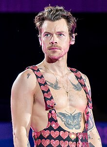 A brown-haired man with light facial hair and a serious expression shows off large bird and butterfly tattoos by being barechested, except for artistic overalls decorated in hearts.