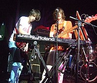 An electric guitarist and a bassist are on stage beside a "JUNO-60 Roland" synthesiser keyboard, which is being played by the guitarist. A drum kit is in the background.