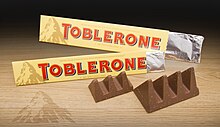 Zwei offene Toblerone-Packungen mit einem groß- und einen kleinzackigem Tobleroneriegel. Zu sehen ist noch das alte Toblerone-Logo in Form des Matterhorns mit einer Schattensilhouette des Berner Bärs auf den Packungen und dem Tisch darunter.