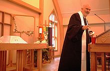 Troy Perry steht in einer Kirche vor einer Kanzel mit einem Mikrofon, in das er reinspricht. Er trägt ein schwarzes Priestergewand und einen grauen Kinnbart. Im Hintergrund sind auf Tischen Pflanzen, eine weiße Kerze in einem Ständer und ein aufgestelltes Holzkruzifix zu sehen. Am Kreuz hängt eine Regenbogenflagge, auf der die Aufschrift Healing Spirit und eine Friedenstaube aufgenäht sind.