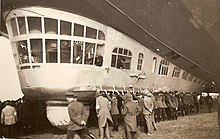 The gondola while the airship is being manoeuvred on the round. Around forty people on the ground are manhandling it. Two officers are visible in the gondola, one looking down at the people, the other looking backwards. The ram air turbine is folded flush with the gondola's side.
