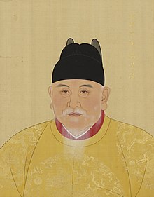 An old aged bearded man wears yellow robes with dragons inscribed and a black hat.