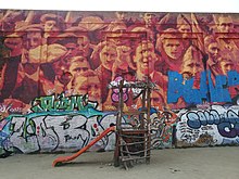 A brick wall depicting a mass of people with raised fists. It has been defaced wit graffiti. A slide for children is in front of it.