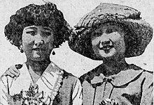 A black-and-white newspaper photograph of two young Asian women, side by side, photographed outdoors. The woman on the left is not smiling or wearing a hat; the woman on the right is smiling and wearing a hat.