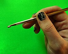 A person with painted nails holds a yellow crochet hook on a bright green background.