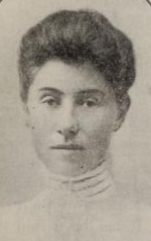 A young white woman with dark hair in a bouffant updo, wearing a white blouse with a high collar