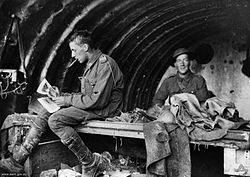 Two men in military uniform in a building. The man on the left has something in his mouth and is sitting down looking at some papers, angled away from the camera. The men on the right is smiling and looking at the camera. The roof of the structure is curved.