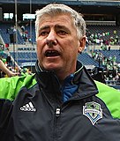 A headshot of Sigi Schmid wearing a Seattle Sounders FC tracksuit, pictured at a stadium