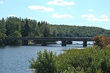 A river flowing between two banks covered in vegetation and trees. The river flows under a concrete bridge resting on four support piers into a lake in the background.