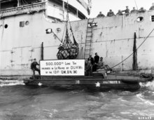 A banner proclaims the 500,000th long ton. It is flanked by Caucasian sailors and stevedores and African-American DUKW drivers.