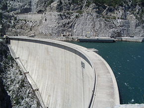 Almost vertical high smooth concrete curving dam in rocky landscape, seen from one side: holding back dark blue water with small waves