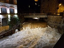 Night shot showing the Durolle in flood, with a very large flow of brownish water at a small waterfall in the middle of town, with a factory on the bank to the left of the shot.