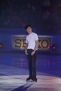 A photograph of Nathan Chen standing on the ice under purple exhibition lights.
