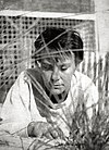 A photograph of Harper Lee in an outdoor setting. She has short hair and appears to be examining something in the sand and grass.