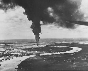 Aerial black and white photo of a river with buildings on its left-hand shore. A large column of smoke is rising from near the bank of the river.