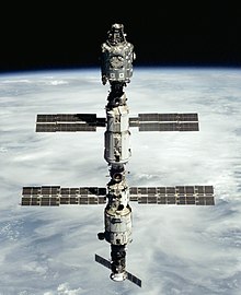 Three modules linked in a linear arrangement float in space with the Earth in the background. The top module is a metallic cylinder with a large white circle visible on it and a black cone at either end. The two lower modules are cylindrical and covered in white insulation, and have two blue solar arrays projecting from each. A smaller, brown spacecraft is docked to the lower module.