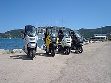 Four BMW C1 scooters parked side by side in front of a lake, with mountains and the masts of a large ship in the background