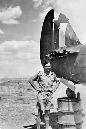 Man in khaki shirt and shorts, wearing forage cap, beside aircraft missing part of tailplane