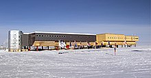 A long, large building consisting of several sections stands behind a line of flags flying on poles. The ground surface is ice-covered; in the middle foreground is a short-striped pole which indicates the position of the South Pole