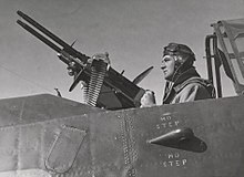 Black and white photograph of a man seated in an aircraft cockpit holding the rear handles of a large pair of guns