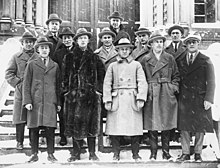 Twelve men pose on the steps in front of a building. They are wearing suits, long jackets and hats.