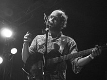 Black-and-white photograph of a bearded man playing guitar onstage.