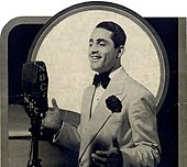 Bowlly, wearing a white dinner jacket and buttonhole, sings into a microphone