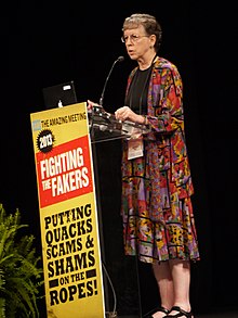 Harriet Hall is standing in front a podium that has the tag lines Fighting the fakers. Putting quacks, scams, and shams on the ropes!