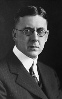 A severe-looking man in round-rimmed glasses
