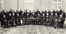 A photograph of a group of men in a semi-circle consisting of two rows. The front row is seated. There is a desk in the middle of the semi-circle with a man seated behind it.
