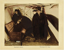An image of a lobby card with a photograph in the center, and small text captions underneath it. The image shows a man in a dark coat, cape, and top hat standing to the right, while two men investigate another man who is sitting upright inside an open box.