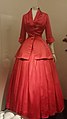 A floor-length red gown. The bodice features a blazer-style jacket with a severe wasp-waist, and the skirt is floor-length, pleated, and bell-shaped.