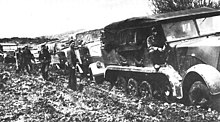 A black and white photograph of a half-tracked prime mover pulling heavy trucks along a muddy road