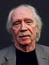 An elderly Caucasian man with a gray mustache and gray receding hair faces the camera with a neutral expression.