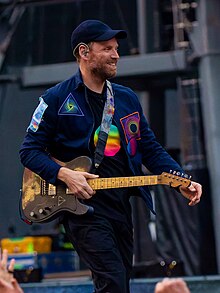 A man wearing a dark blue cap and jacket walks with his guitar